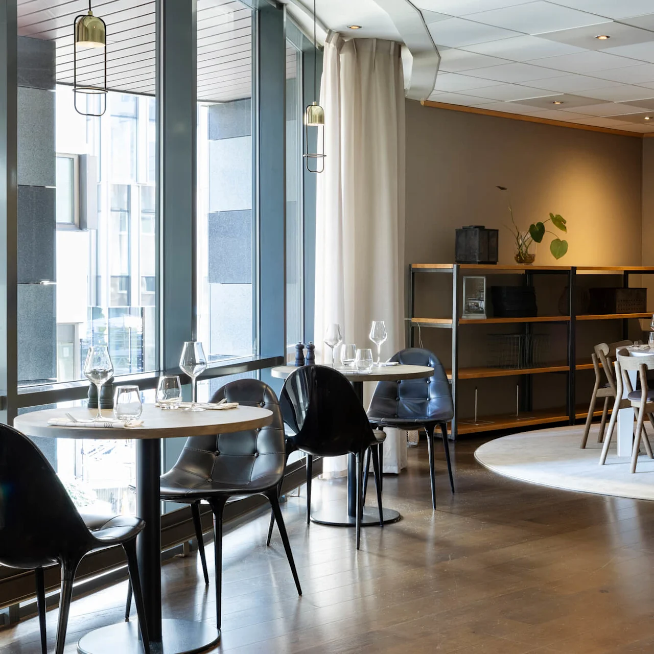 A dining table, set with glassware and cutlery, occupies a modern restaurant with wooden floors, black chairs by large windows, and soft lighting. 