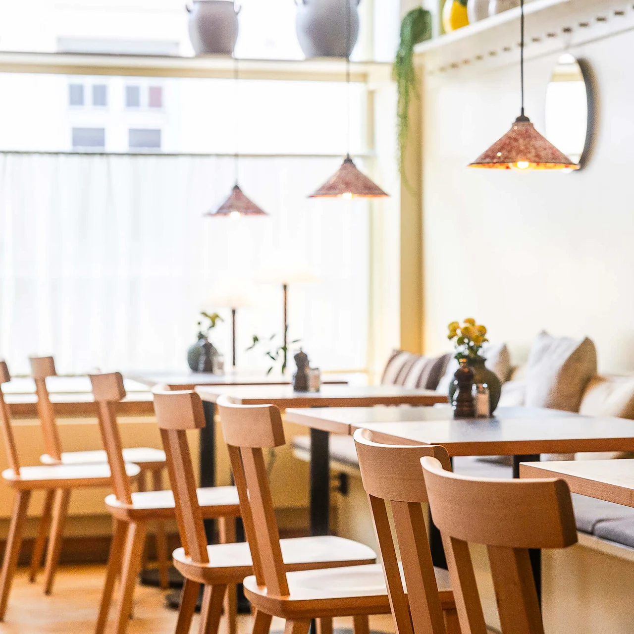 Trästolar och bord i ett mysigt, välbelyst kafé med hängande lampor och växter vid stora fönster.