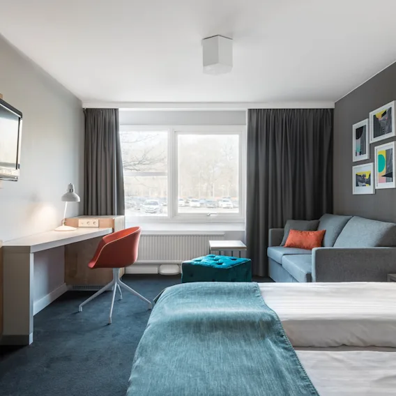 Ett hotellrum med en säng täckt av turkost sängkläder, en väggmonterad TV, en orange stol vid ett skrivbord och en grå soffa vid ett fönster med utsikt över parkerade bilar.