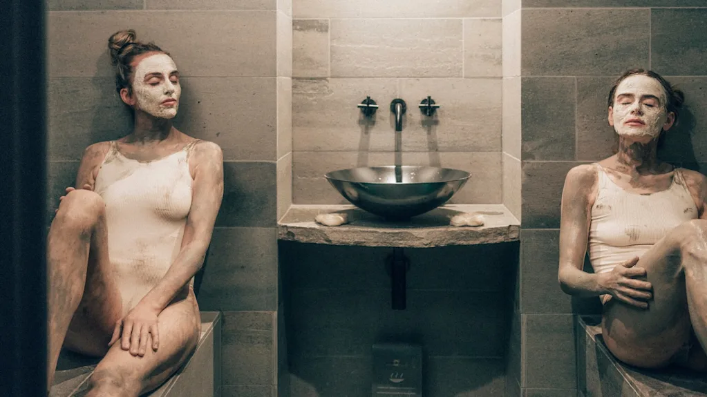 Two people with mud masks sit relaxed, wearing swimsuits, in a dimly lit spa area. A stone basin and wall-mounted faucets are centered between them.