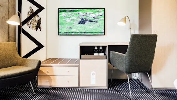 Et vægmonteret TV viser en naturscene i et moderne rum med to lænestole, et skrivebord, et skab og dekorativ kunst. Tekst på TV: "UTVAN?"