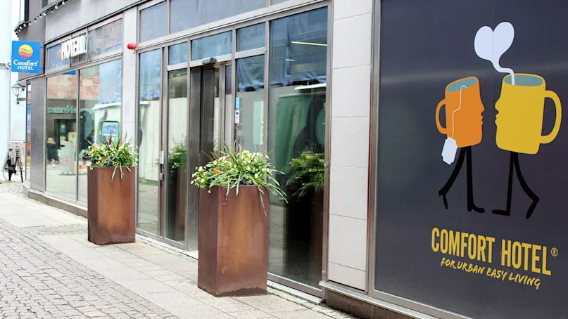 Two stylized mugs form faces; one has a steam heart above. "COMFORT HOTEL - FOR URBAN EASY LIVING" is written. It's outside, with plants and a cobbled street.