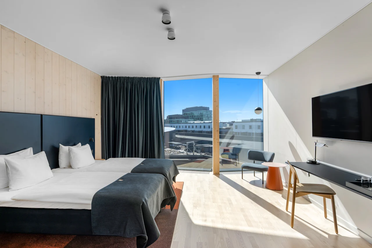 Et lyst, moderne hotelværelse med to enkeltsenge, et stort vindue med udsigt over byen og et behageligt siddeområde.