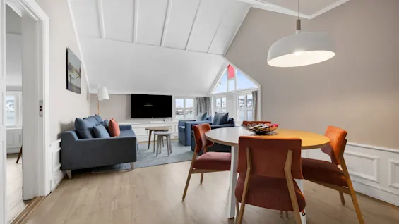 Stuen har en blå sofa vendt mot en veggmontert TV under høyt, hvitt tak. Et trebord med røde stoler og en stor pendellampe komplementerer den koselige moderne innredningen.