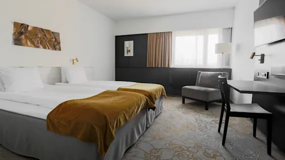 A neatly made bed with brown throws, sits in a minimalistic hotel room with white walls, a gray armchair, and a black desk next to a large window.