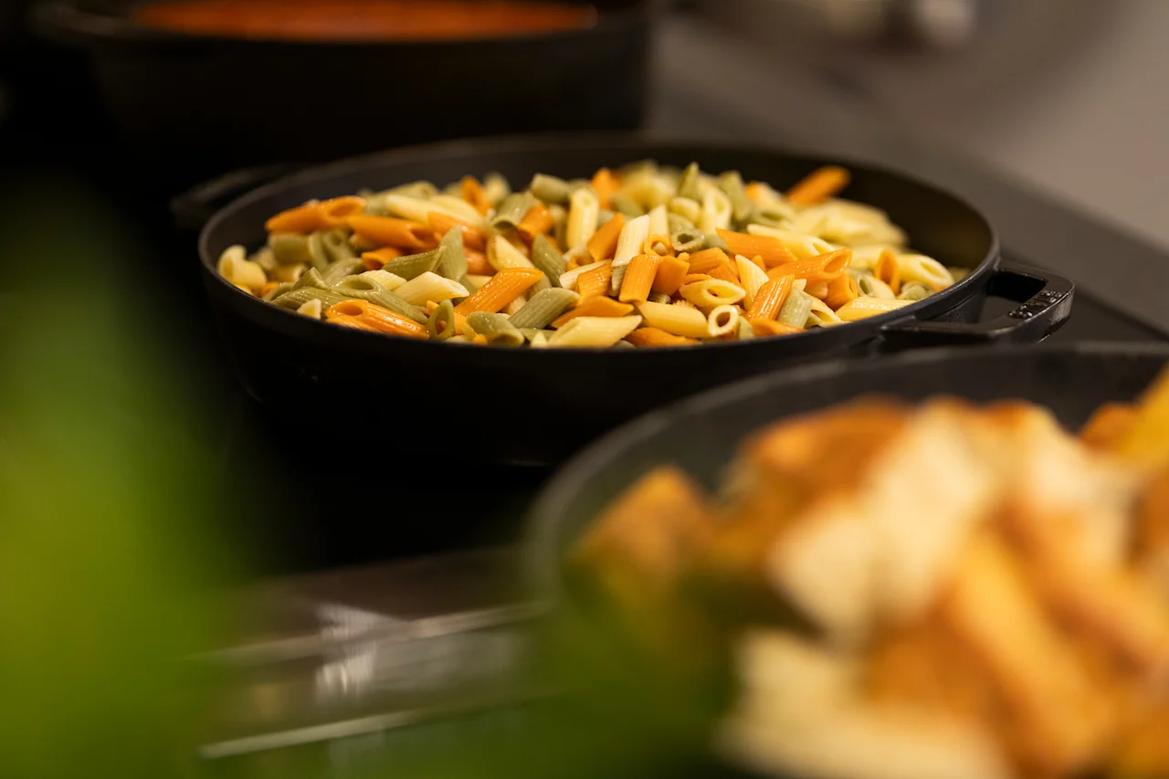 A close-up of a serving dish filled with colorful penne pasta, suggesting a delicious meal ready to be enjoyed at a buffet.