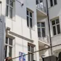 En gård har en sitthörna med stolar och bord, omgiven av en vit flerbostadsbyggnad med fönster. Solljus kastar skuggor och ett färgglatt konstverk syns på väggen.