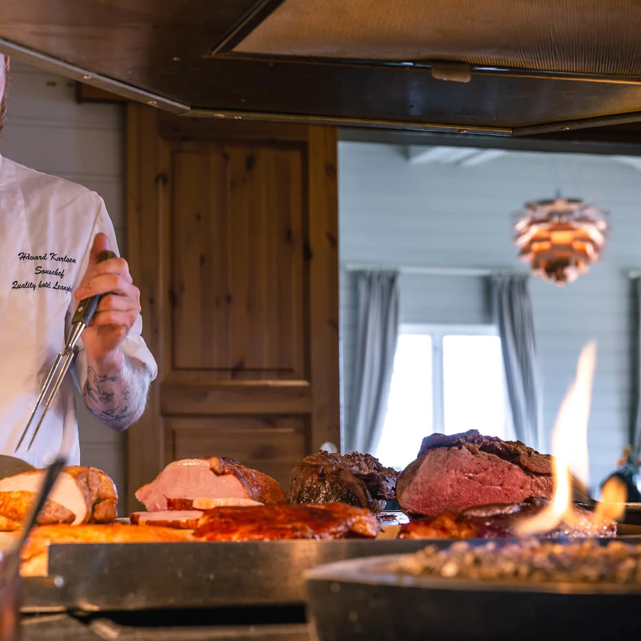 Kokk i hvit uniform skjærer stekt kjøtt med kniv og gaffel i et varmt belyst kjøkken. Tekst på jakken lyder "Harald Karlsson" og "Sous-chef Quality Hotel Leangkollen."