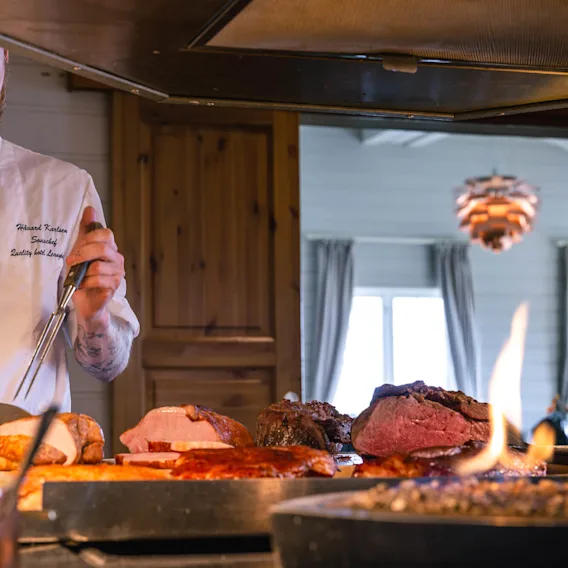 Kokk i hvit uniform skjærer stekt kjøtt med kniv og gaffel i et varmt belyst kjøkken. Tekst på jakken lyder "Harald Karlsson" og "Sous-chef Quality Hotel Leangkollen."