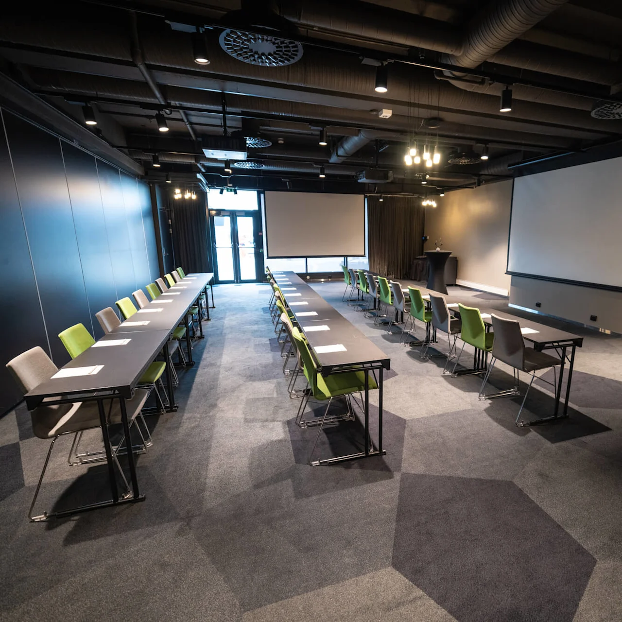 Et konferencelokale med arrangerede borde og stole, der danner en midtergang, under et industrielt loft. To store projektionsskærme er foran.