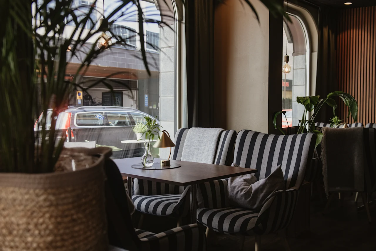 Randiga stolar omger ett litet bord med en växt och lampa i ett mysigt kafé. Stora fönster visar parkerade bilar utanför, och krukväxter pryder interiören.