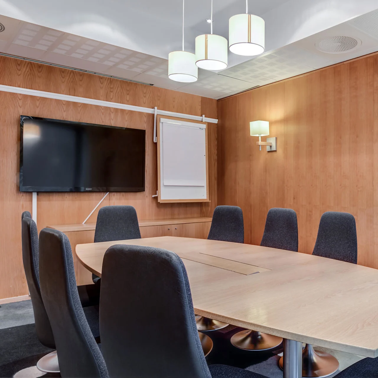 A conference table surrounded by black chairs sits under hanging lamps in a wood-paneled meeting room, equipped with a wall-mounted TV and a flip chart.