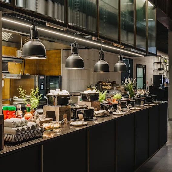 A spacious buffet counter in a modern restaurant is laden with black pots, dishes, eggs, herbs, and condiments. Overhead, industrial-style pendant lights provide illumination.