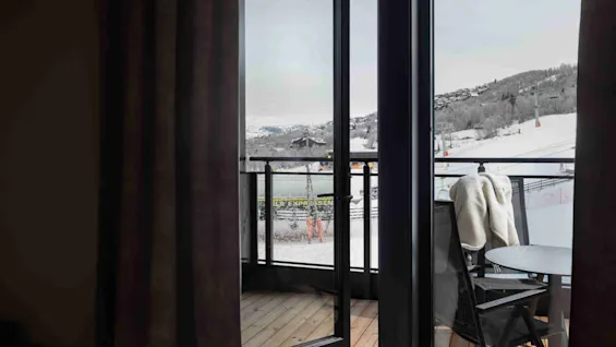 En kop står på en seng nær en altandør. Den åbne dør afslører et snedækket landskab med bakker og skiløjper. En stol og et bord er på altanen.