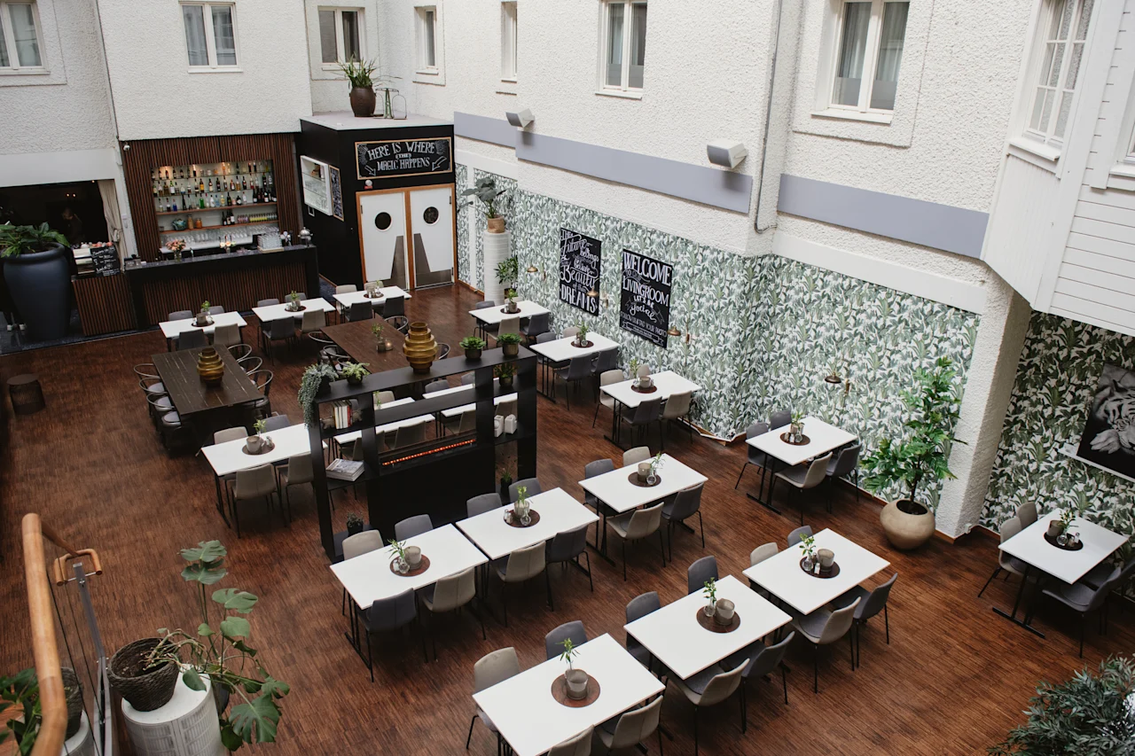 En matsal med prydligt arrangerade bord och stolar, dekorerade med krukväxter, ligger i ett inomhusområde med växtmålningar och en bar. Skyltar säger "Limewood Bar & Restaurant," "Restaurant Enters," och "Welcome Open."