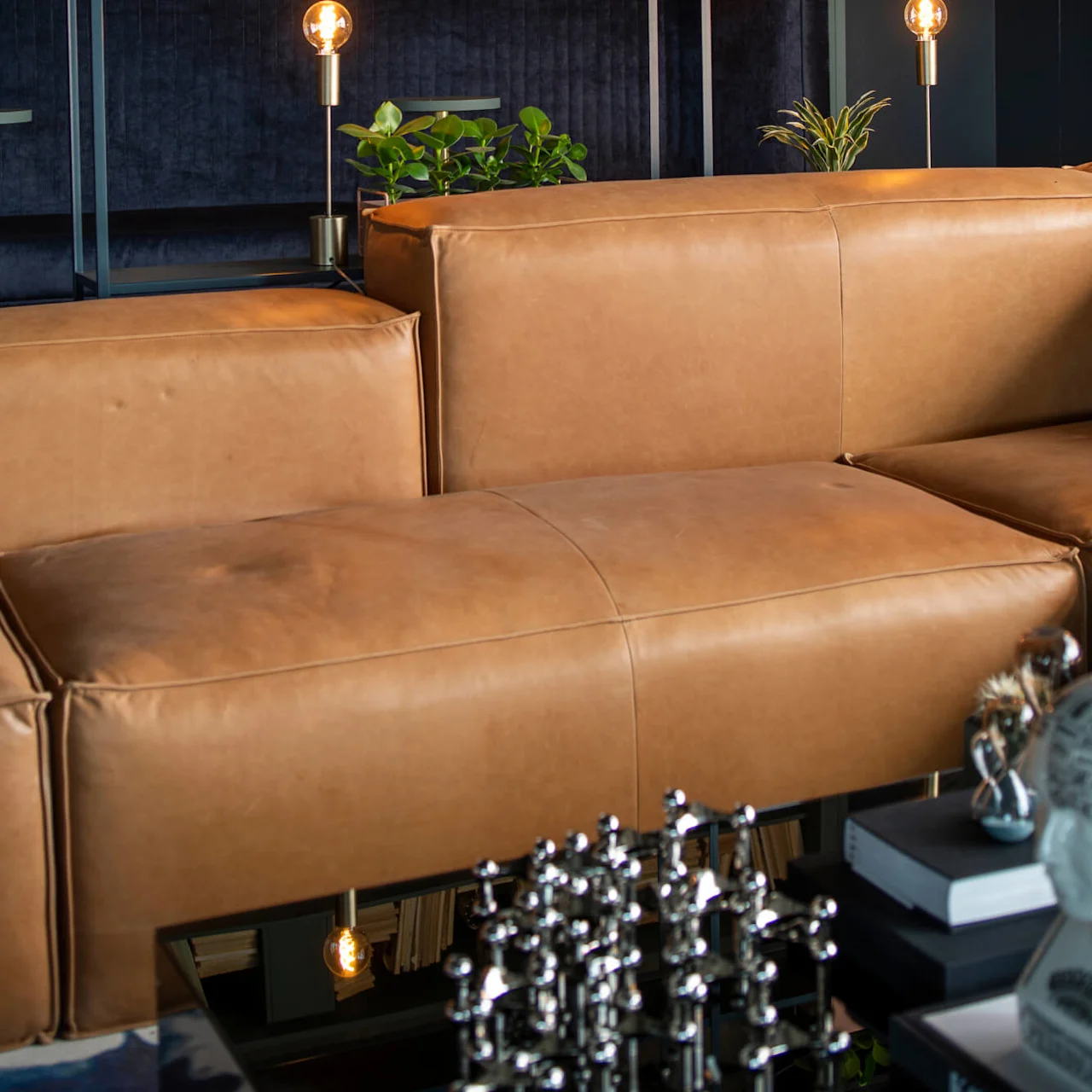 A brown leather sofa sits in a modern living room, accented by plants, bookshelves, and soft lighting. The environment is cozy with a glass table and decorative items.