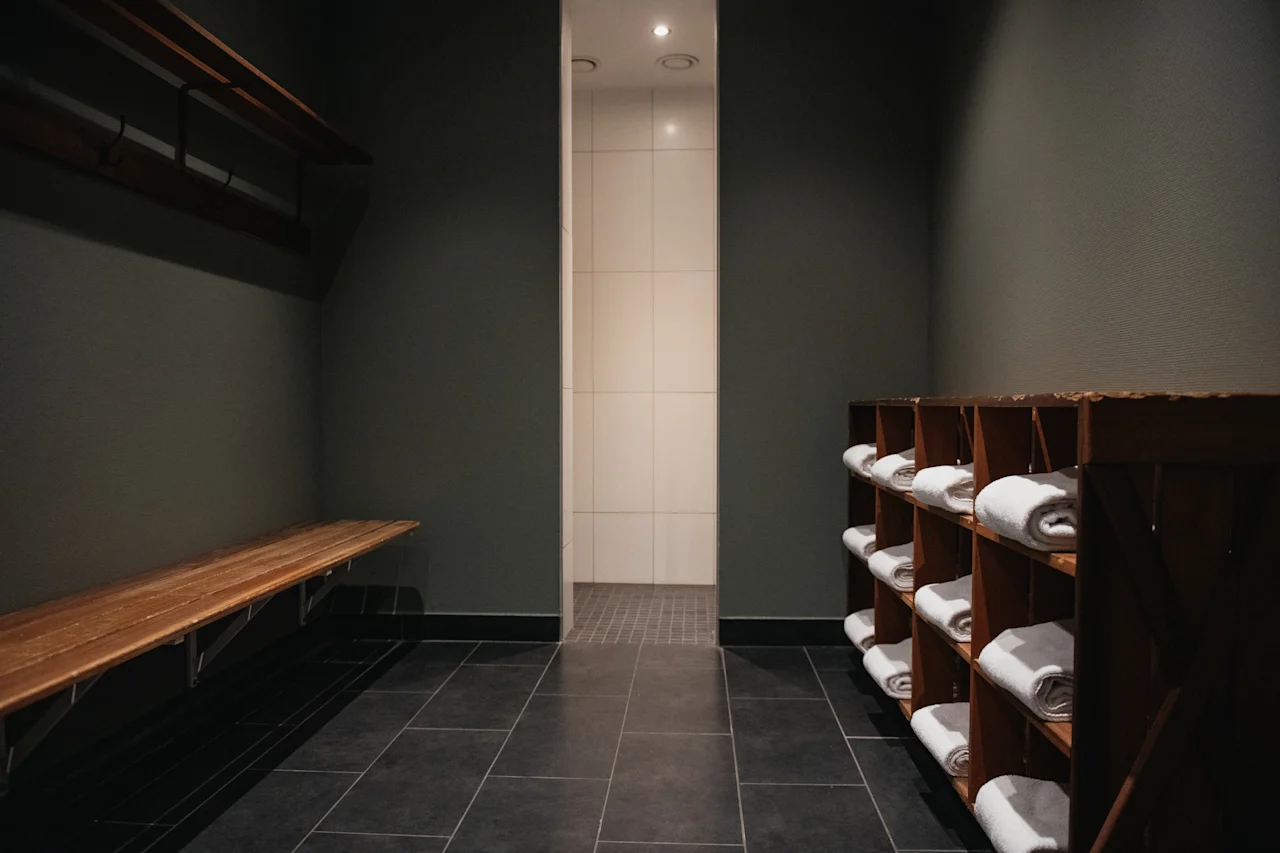 Ett lugnt omklädningsrum med en träbänk och fräscha vita handdukar, som leder till ett ljust duschutrymme.