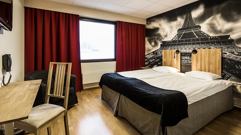 Ett hotellrum med två enkelsängar, mörkröda gardiner, en stor väggmålning av Eiffeltornet och enkel inredning som föreslår komfort och modern interiör.