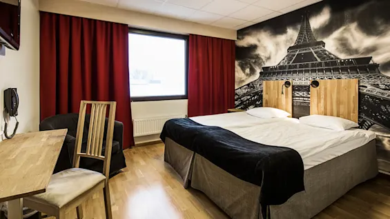 A hotel room with twin beds, dark red curtains, a large mural of the Eiffel Tower, and simple furniture, suggesting comfort and modern interiors.