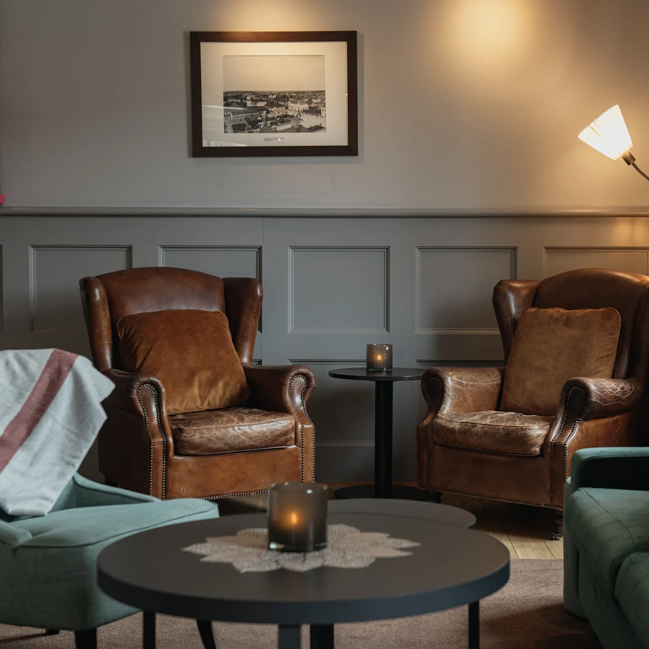 Two leather armchairs sit facing a small round table with candles in a cozy living room. Nearby, a lamp and bookshelves add warmth to the paneled walls.