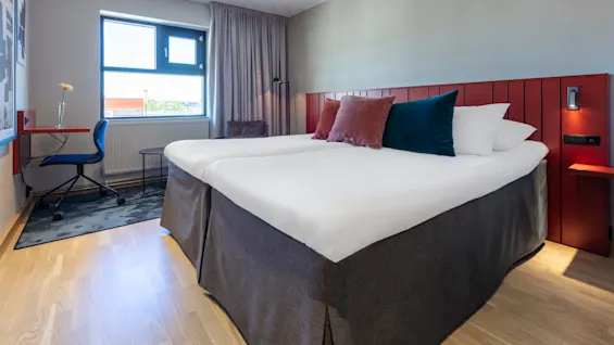 A hotel room with a neatly made bed, a desk and a chair, illuminated by natural light from a window.