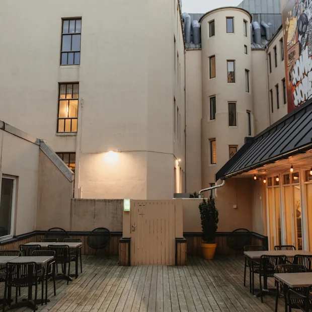 En indbydende udendørs terrasse med trægulv, borde og stole, omgivet af bygninger med varme lys og vægmaleri.