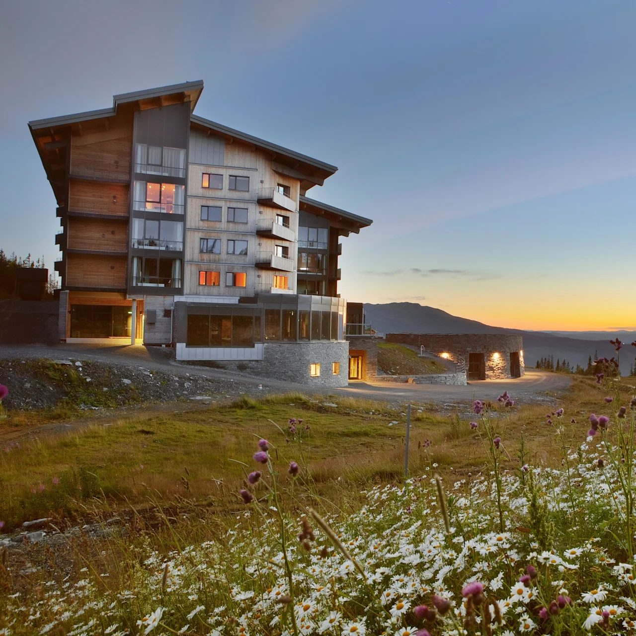 A modern building at dusk with illuminated windows, surrounded by wildflowers, in a mountainous landscape.