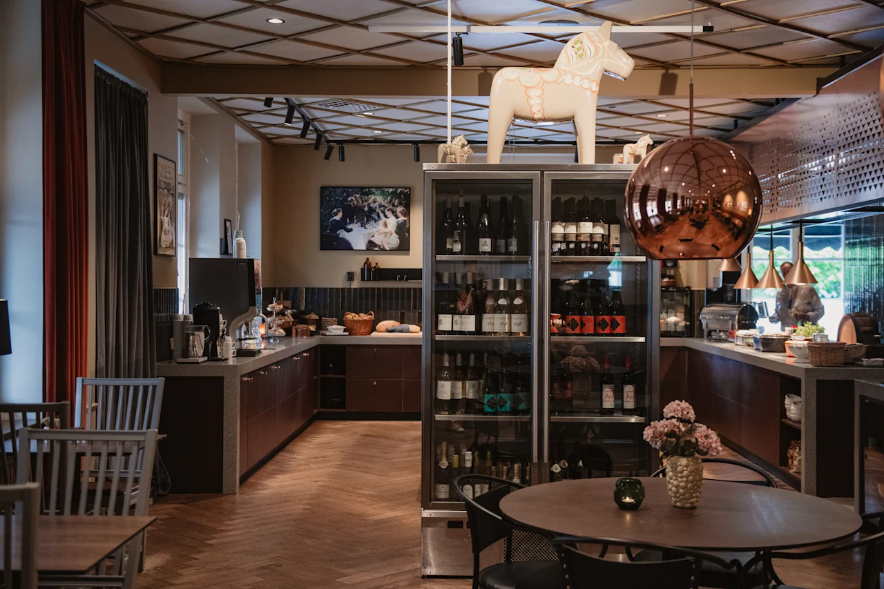 Et koselig hotellounge- eller restaurantområde med en selvbetjeningsdisk, et kjøleskap og sitteplasser. En dekorativ hest står oppå kjøleskapet.