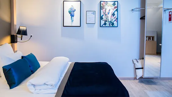Seng med hvide lagner og blå puder placeret i et moderne soveværelse, udsmykket med indrammet kunst på væggen og et spejl i fuld længde ved siden af et rummeligt trægulv.