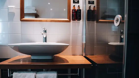 En hvid håndvask med en sølvhane står på en træbordplade. To håndklæder hænger under, og toiletartikeldispensere er monteret på den flisebeklædte væg nær et spejl.