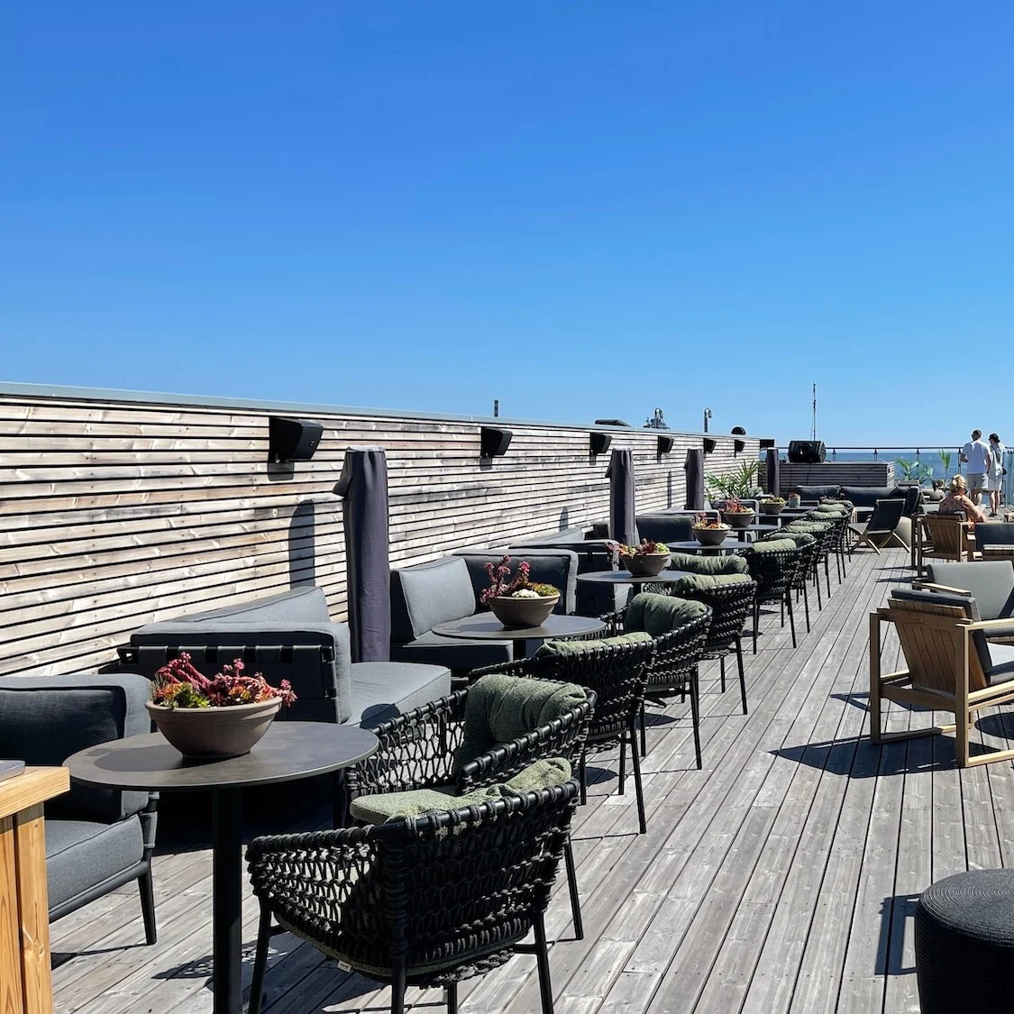 En tagterrasse med moderne møbler, der ser ud over et stille vand, under en klar blå himmel.