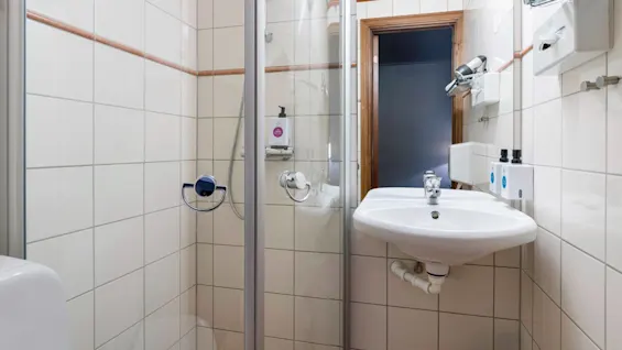 Brusekabine med glasdør og håndholdt bruser i et flisebelagt badeværelse, ved siden af en vægmonteret håndvask med tandpasta dispensere. Håndklæder og toiletskab er synligt.