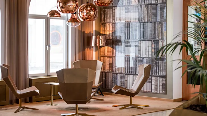 Fire moderne stoler omgir et lite bord under kobberlamper i et rom med en vegg designet som bokhyller. Store vinduer og planter skaper en innbydende atmosfære.