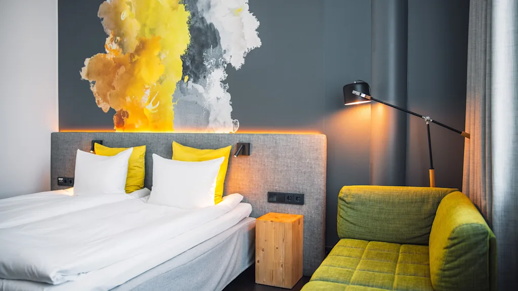 A modern hotel room features a neatly made bed with white and yellow pillows, a vibrant abstract wall painting, a green sofa, and a wall-mounted reading lamp, creating a cozy ambiance.