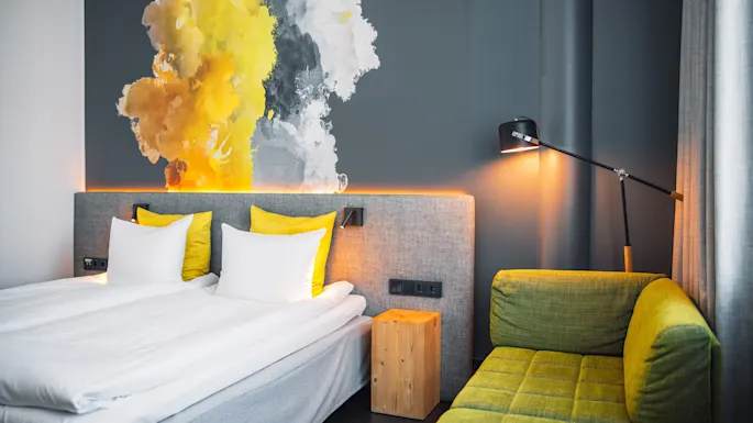Moderni hotellihuone, jossa on siististi pedattu sänky valkoisilla ja keltaisilla tyynyillä, elävä abstrakti seinämaalaus, vihreä sohva ja seinään kiinnitetty lukuvalaisin luovat viihtyisän tunnelman.