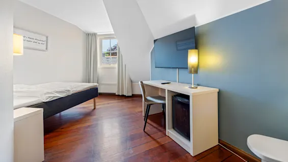 A tidy bedroom with a single bed, desk, lamp, and TV, set against blue and white walls suggesting calmness.