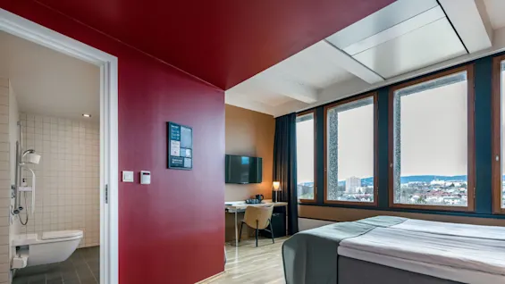 A neatly arranged hotel room with a bed, desk, and TV. Large windows overlook a cityscape. Adjacent bathroom has tiled walls. Walls are red, and floor is wooden.