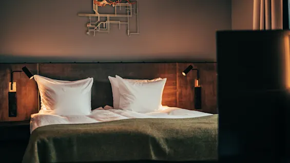 A cozy hotel room with a neatly made bed, white pillows, and a green blanket, illuminated by warm bedside lamps.