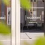 Glasdøre viser teksten "TOLLBODEN HOME HOTEL" og reflekterer en gade med bygninger. Grønne blade indrammer delvist udsigten, hvilket indikerer en udendørs setting.