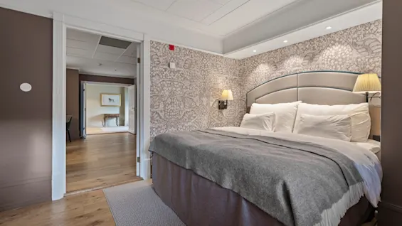 A welcoming hotel room with a neatly made bed, decorative wallpaper, and an open doorway revealing an adjacent space.