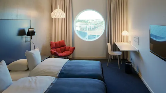 Two neatly made beds face a wall-mounted TV in a modern hotel room. A red chair, a circular window, and a small desk with a lamp complete the minimalist decor.