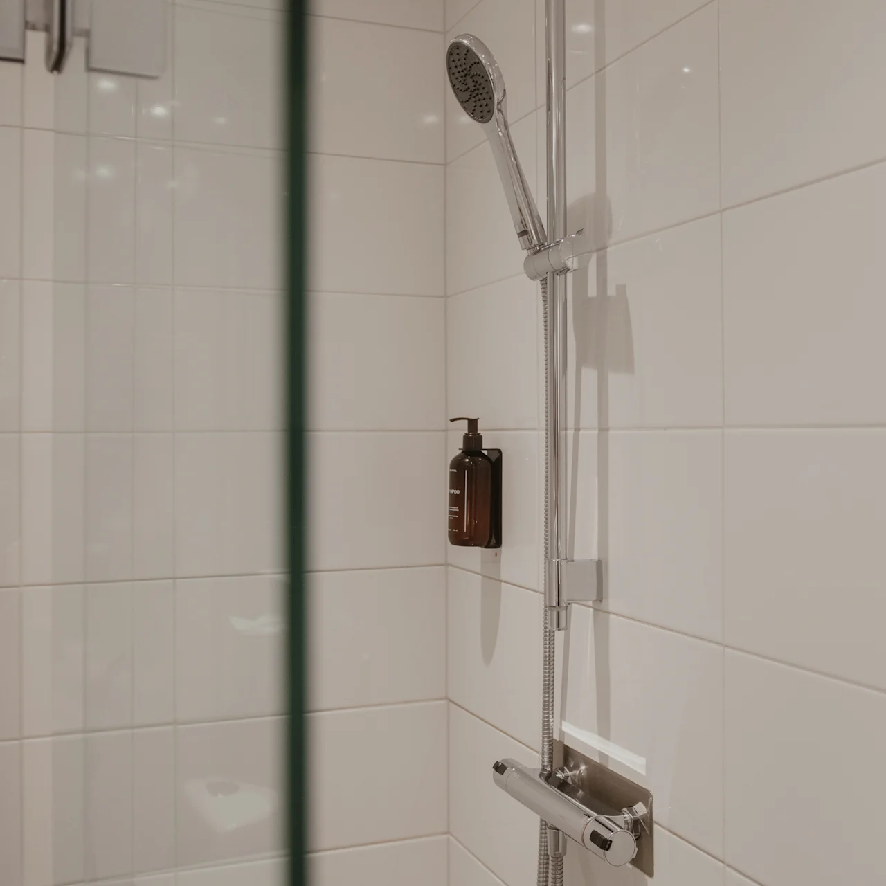 Brusehoved fastgjort til flisevæg, oven over en lille brun flaske; lukket af en klar glasplade i et moderne badeværelse.