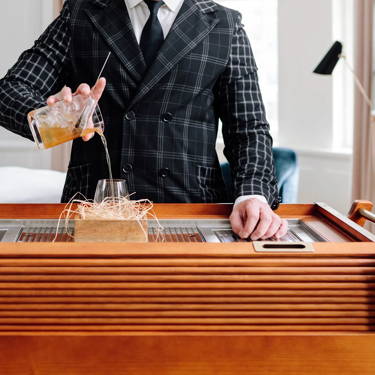 A person in a checkered suit pours a drink into a glass on a wooden cart in a room with a bed and lamp.