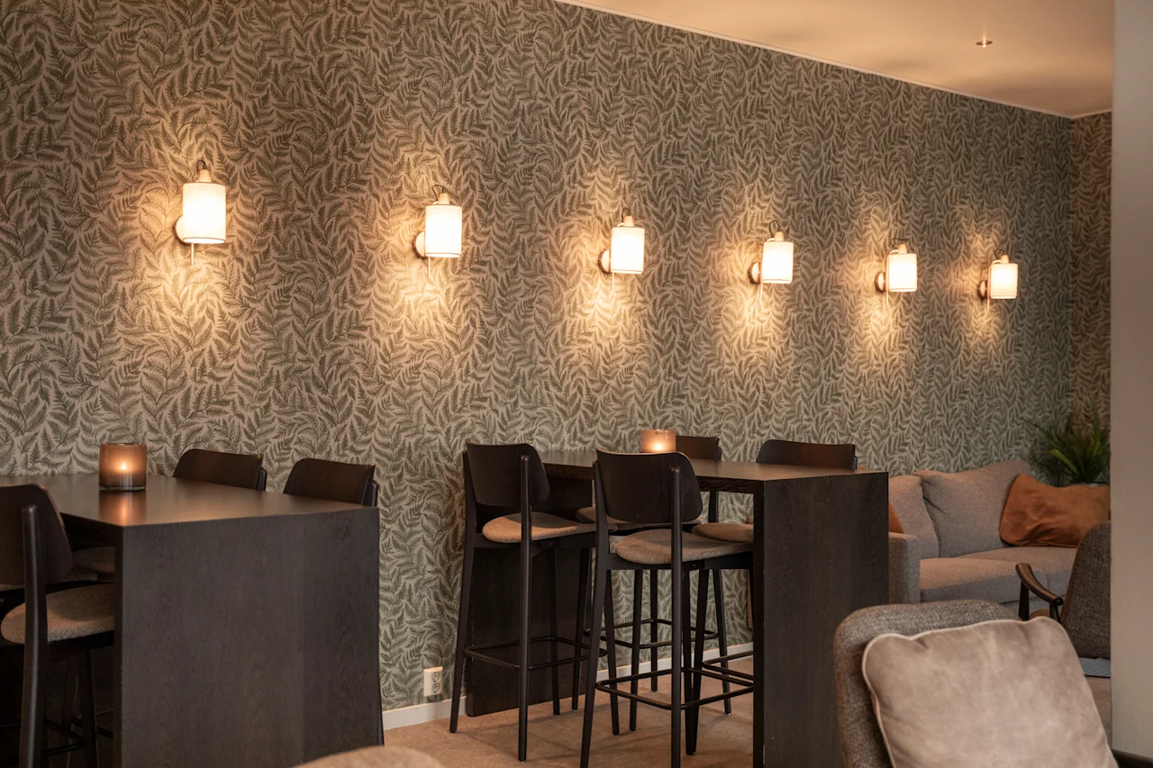 Viihtyisä oleskelutila, jossa on kuviollinen tapetti, lämmin seinävalaistus, korkeat pöydät jakkaroineen ja mukavat sohvat.