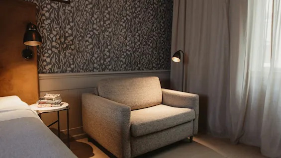 A cozy hotel room with a comfortable armchair, patterned wallpaper, and soft lighting, inviting relaxation.