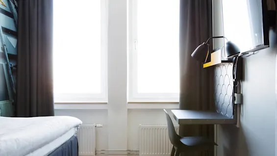 En säng med vita sängkläder står bredvid två stora fönster, intill ett litet skrivbord med en svart stol och lampa. Rummet har trägolv och vita väggar.