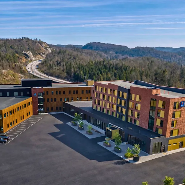 En moderne hotellbygning med en stor parkeringsplass, mot et bakteppe av skogkledde åser og en svingete vei.