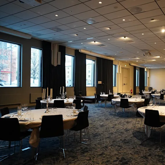Round tables set with elegant dinnerware and surrounded by black chairs in a spacious, carpeted banquet hall with large windows and ceiling lights.