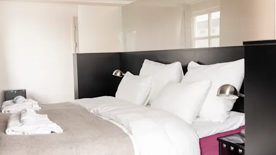 Mukava hotellisänky raikkailla valkoisilla liinavaatteilla ja pehmeällä harmaalla peitolla, kutsuen vieraat rentoutumaan. 🛏️✨