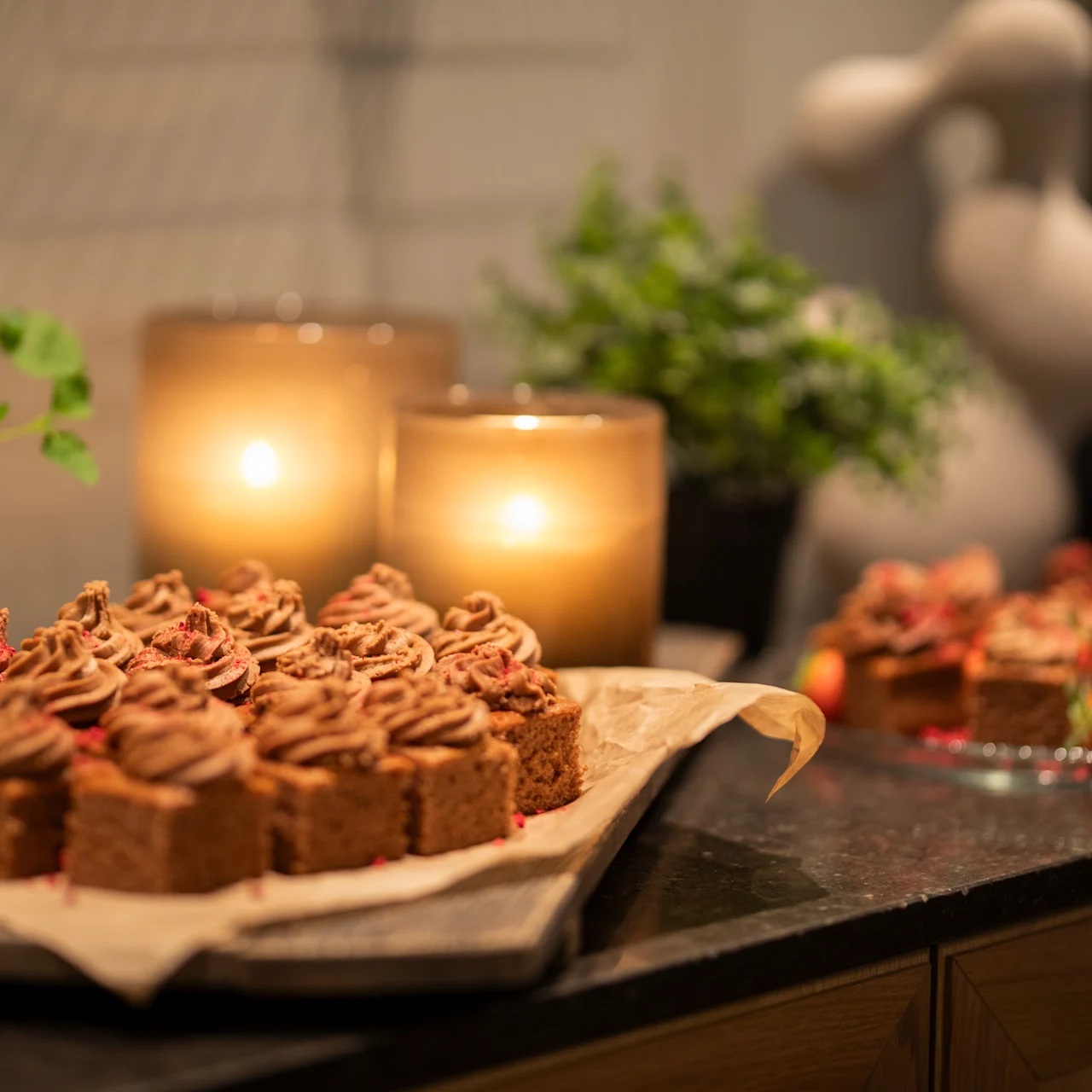 Chokladmuffins med virvlad glasyr ligger på ett fat. Två tända ljus och krukväxter dekorerar den mörka ytan och skapar en varm, inbjudande atmosfär.